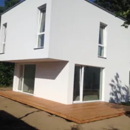 Nowoczesny dom z białym tynkiem i drewnianym tarasem, widoczne okna z białymi ramami i cień drzewa na ścianie.