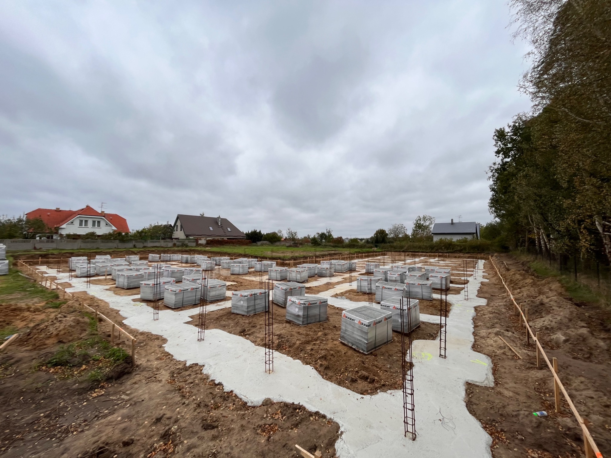 Przygotowany teren pod budowę domu z wylanymi fundamentami i zbrojeniami, na których ułożone są bloczki fundamentowe Durabet w opakowaniach foliowych, widoczne drewniane szalunki, w tle zabudowa...