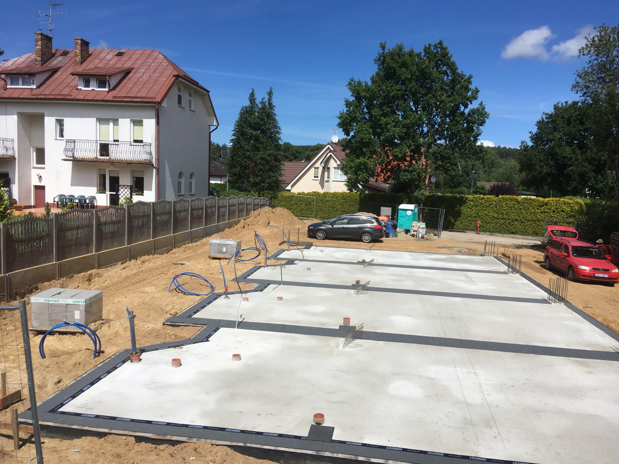 Wylany fundament z izolacją, przygotowany pod budowę domu, widoczne wystające pręty zbrojeniowe, rury instalacyjne i paleta z bloczkami Ginter na placu budowy w słoneczny dzień.