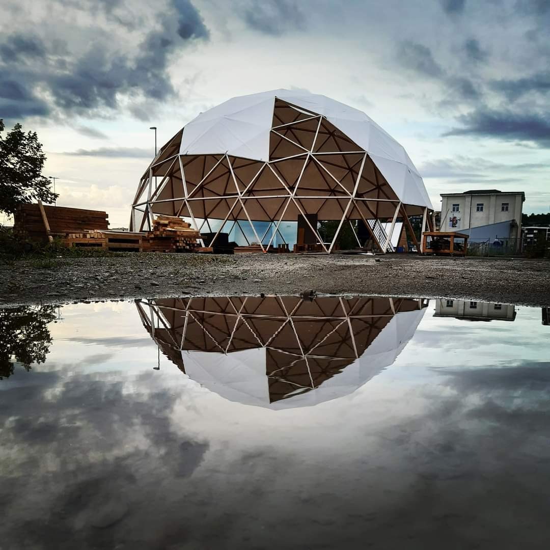 Geodezyjna kopuła z drewnianą konstrukcją i białym pokryciem, częściowo w budowie, odbijająca się w kałuży na pierwszym planie, z paletami i drewnem obok.