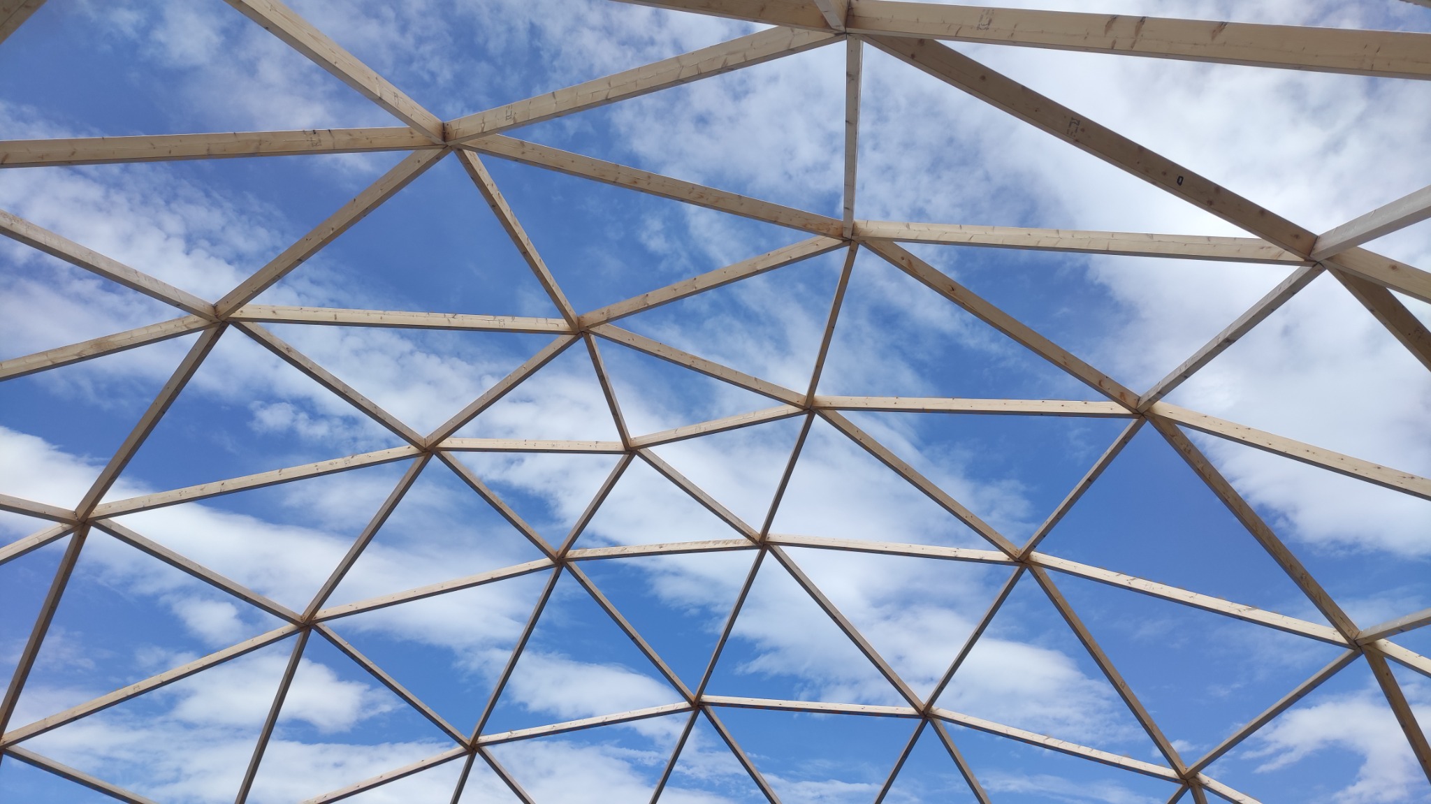 Konstrukcja drewnianej altany geodezyjnej w trakcie budowy, widok od dołu na niebieskie niebo z chmurami.