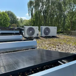 Dwa klimatyzatory Midea i Hoysun na dachu pokrytym kamieniami i mchem, obok paneli słonecznych, w tle drzewa i budynek. Słoneczny dzień.