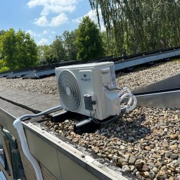 KZ Instalacje Techniczne - Biała jednostka zewnętrzna klimatyzacji Kaysun zamontowana na płaskim dachu pokrytym kamieniami, z widocznymi panelami słonecznymi w tle i fragmentem rynny.