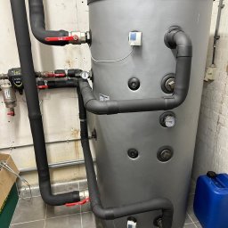 KZ Instalacje Techniczne - Szary zbiornik akumulacyjny z czarnymi rurami, manometrami i zaworami w pomieszczeniu technicznym. Widoczny filtr i niebieski kanister.