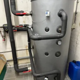 Szary zbiornik akumulacyjny z czarnymi rurami, manometrami i zaworami w pomieszczeniu technicznym. Widoczny filtr i niebieski kanister.