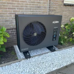 Nowoczesna, szara pompa ciepła Alavo Inverbost na tle ceglanej ściany i kwitnących hortensji, otoczona białymi kamykami.