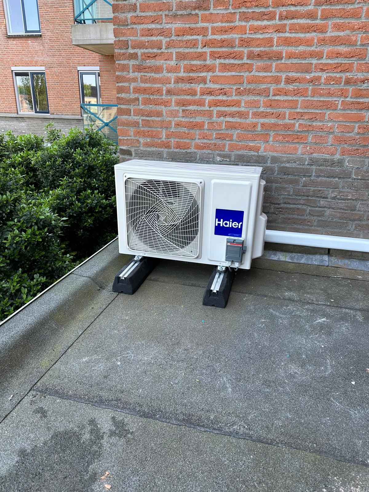 Biała jednostka zewnętrzna klimatyzacji Haier zamontowana na dachu, widok z boku, na tle ceglanego budynku i zieleni.