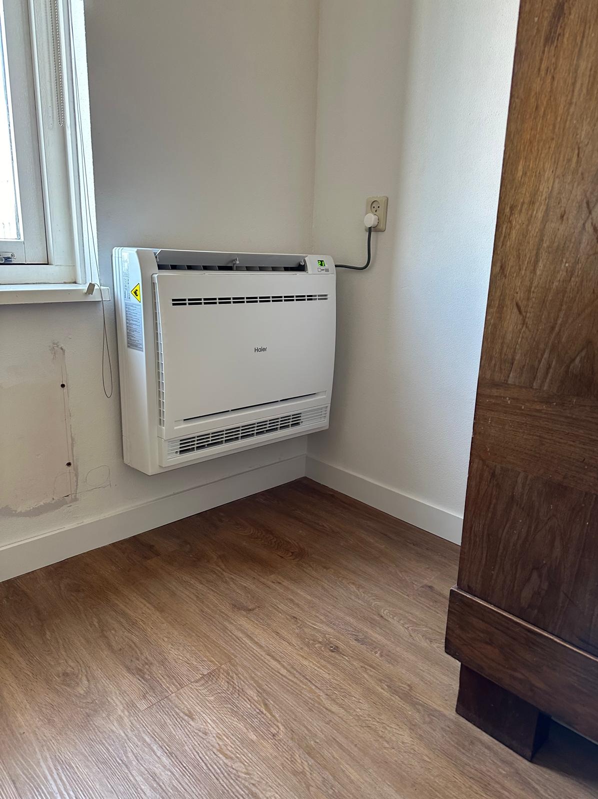 Biały klimatyzator Haier zamontowany na ścianie w rogu pokoju, widoczny fragment okna i drewnianych drzwi, brązowe panele podłogowe.