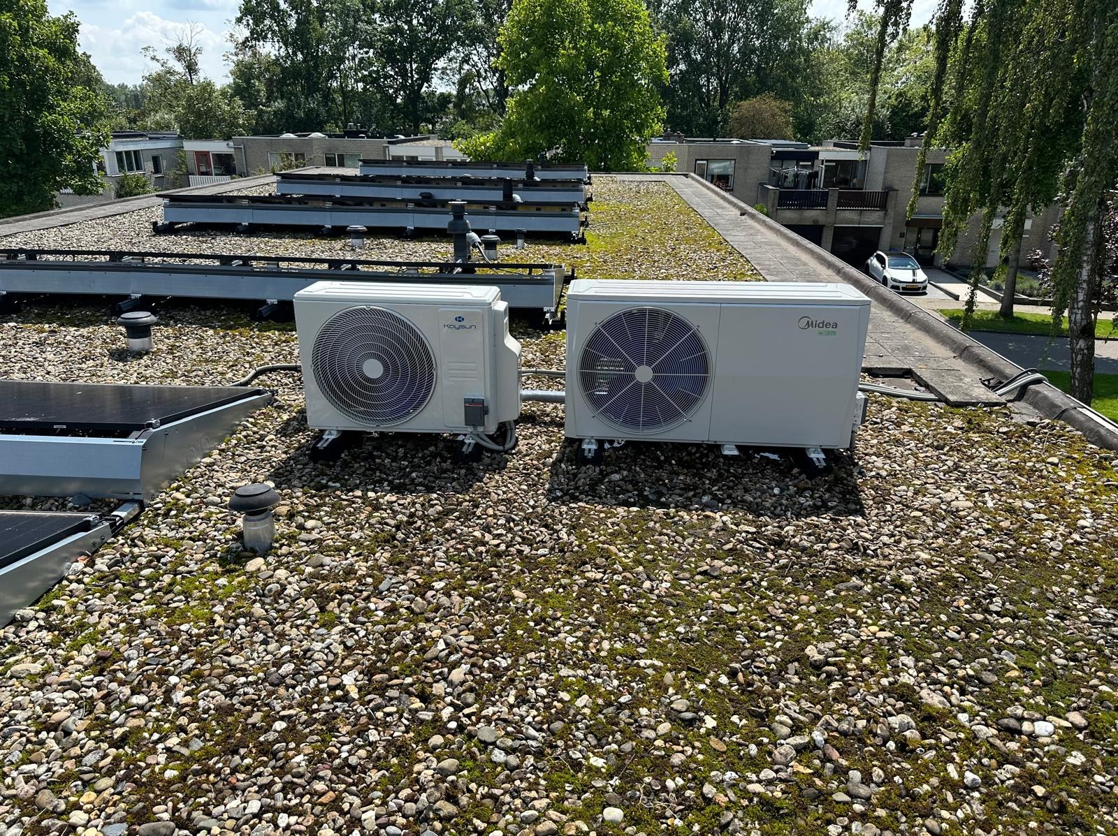 Dwa klimatyzatory (Kaysun i Midea) na dachu pokrytym kamieniami i mchem, z widocznymi panelami solarnymi w tle, w słoneczny dzień.