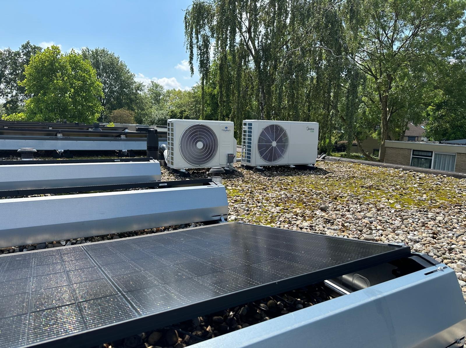 Dwa klimatyzatory Midea i Hoysun na dachu pokrytym kamieniami i mchem, obok paneli słonecznych, w tle drzewa i budynek. Słoneczny dzień.