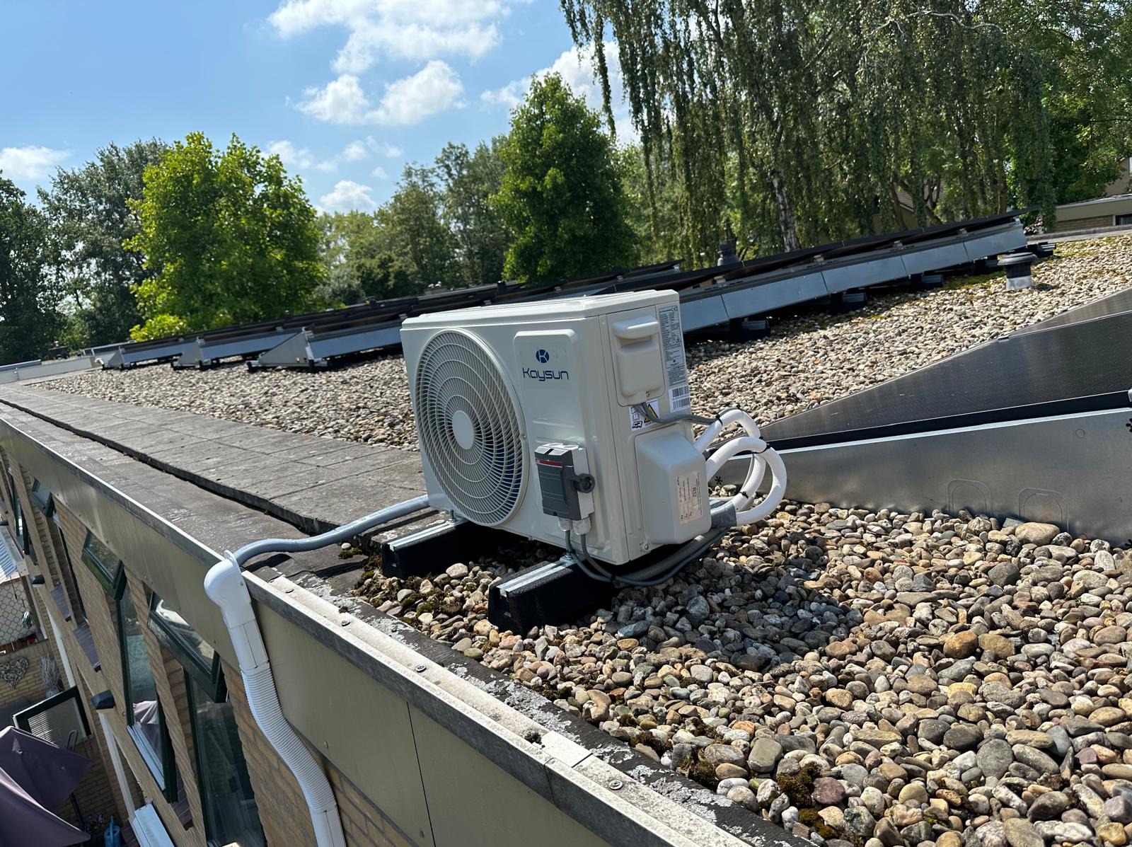Biała jednostka zewnętrzna klimatyzacji Kaysun zamontowana na płaskim dachu pokrytym kamieniami, z widocznymi panelami słonecznymi w tle i fragmentem rynny.