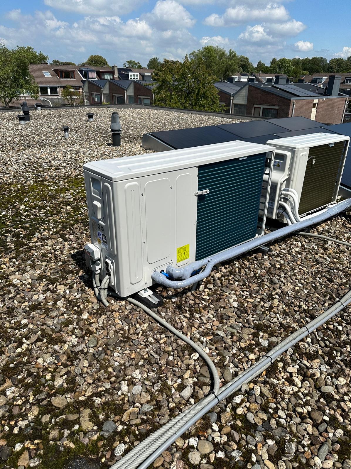 Dwie jednostki zewnętrzne pompy ciepła na dachu pokrytym kamieniami, z widocznymi panelami słonecznymi w tle i budynkami mieszkalnymi.