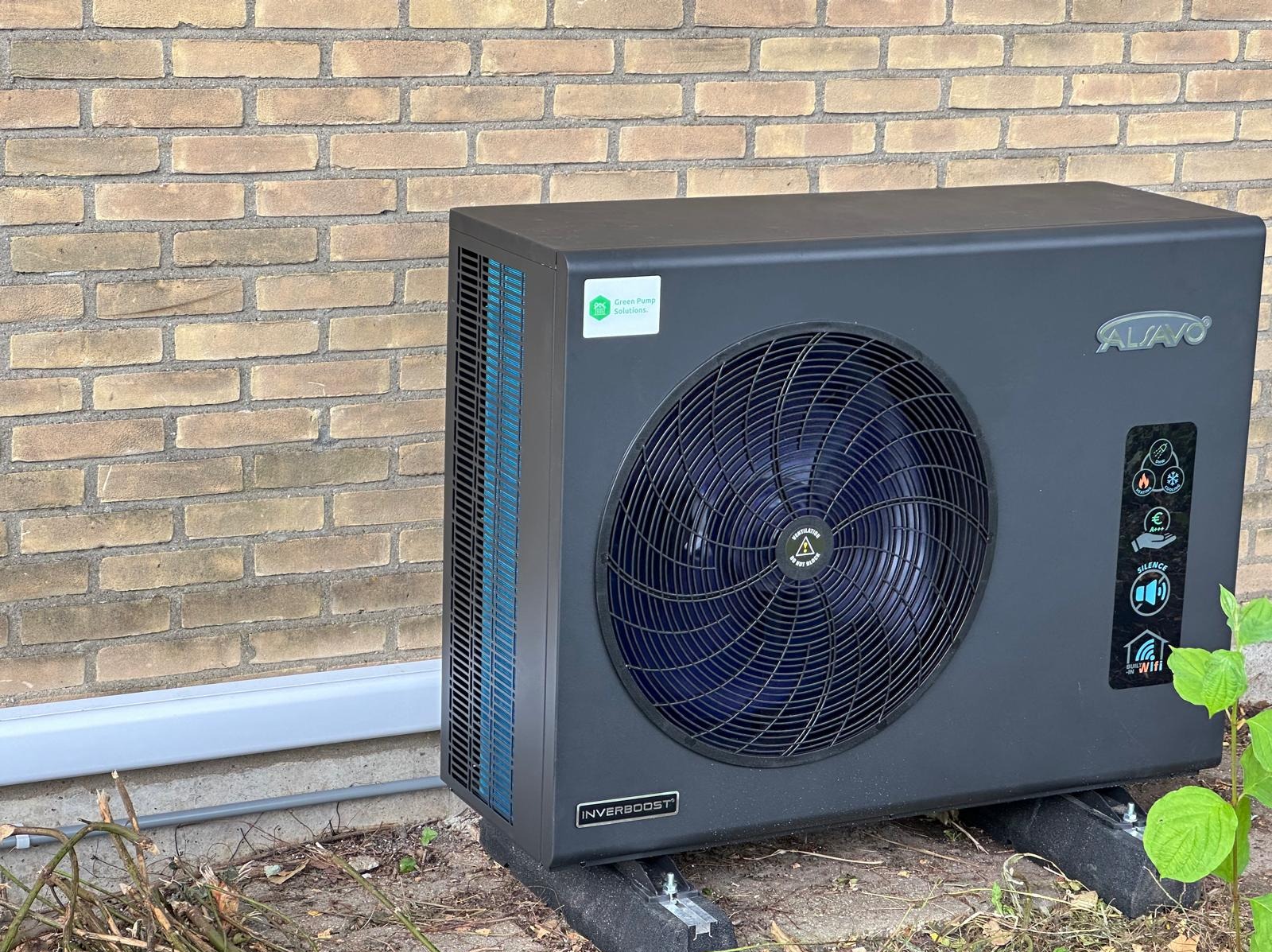 Czarna pompa ciepła Alavo Inverbost, zamontowana na zewnątrz budynku z cegły, z widocznymi elementami instalacji i zielenią. Widoczne oznaczenia i logo producenta.