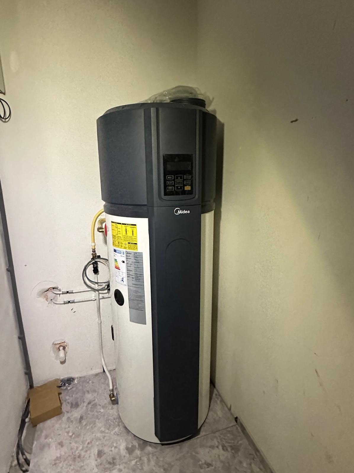 Stojąca pompa ciepła Midea z podłączonymi przewodami w rogu pomieszczenia. Widoczne ślady po instalacji na ścianie. Oświetlenie sufitowe.