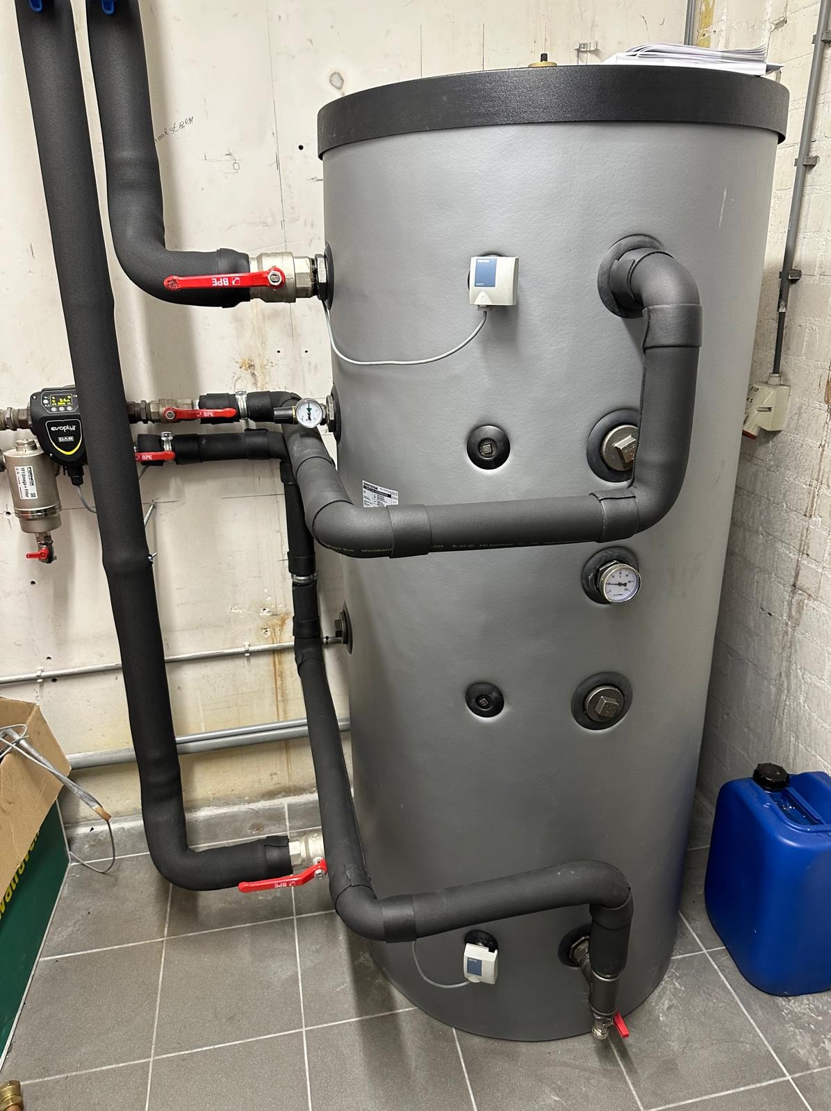 Szary zbiornik akumulacyjny z czarnymi rurami, manometrami i zaworami w pomieszczeniu technicznym. Widoczny filtr i niebieski kanister.