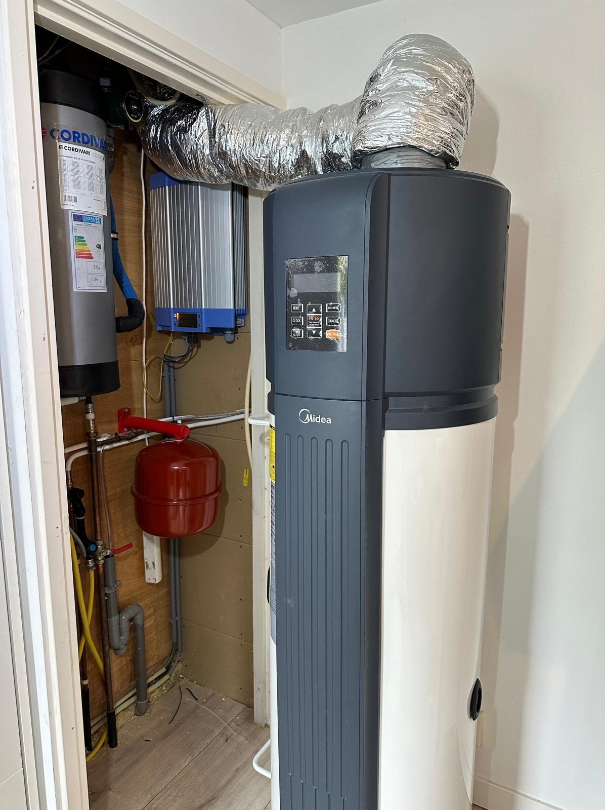 Nowoczesna pompa ciepła Midea w kotłowni, z widoczną instalacją i zbiornikiem Cordivari. Srebrny przewód wentylacyjny łączy system.
