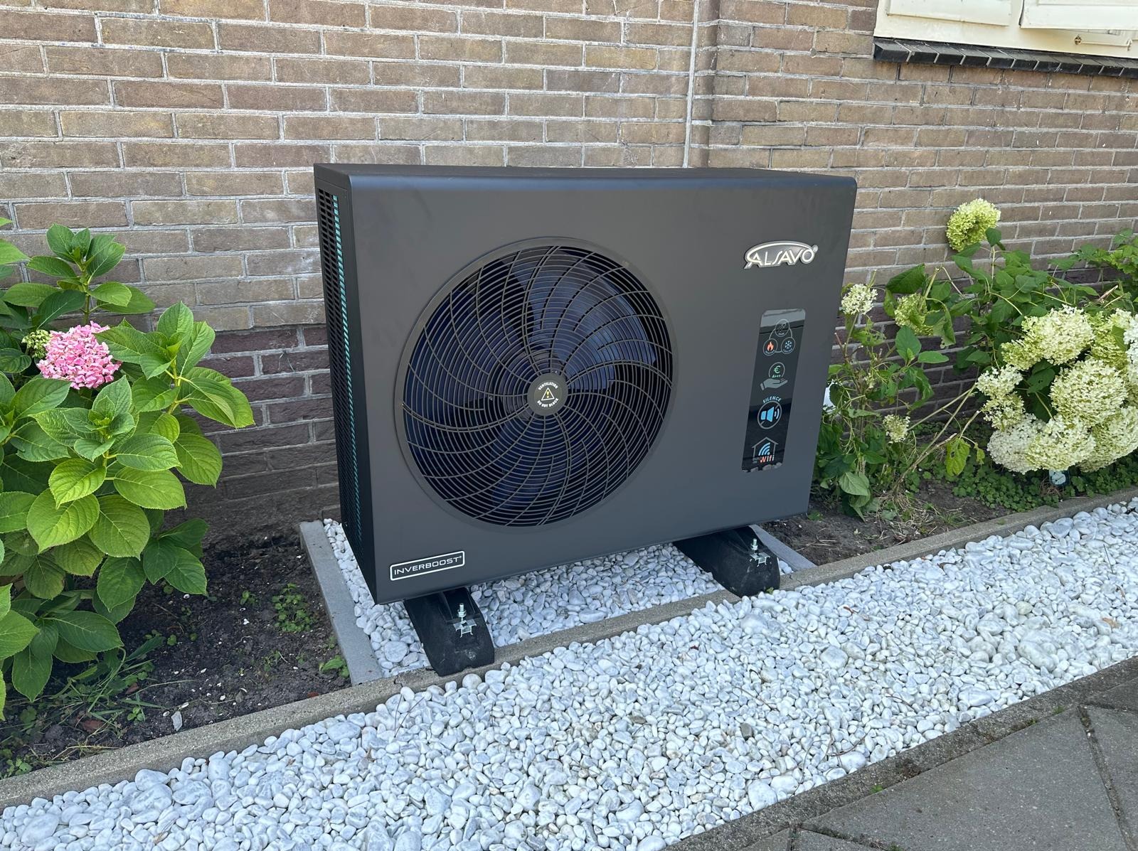 Nowoczesna, szara pompa ciepła Alavo Inverbost na tle ceglanej ściany i kwitnących hortensji, otoczona białymi kamykami.