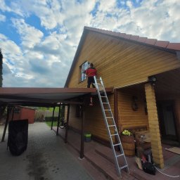 DMM Klimat Dawid Matejek - Instalator na drabinie montuje zewnętrzną jednostkę klimatyzacji na drewnianej ścianie domu jednorodzinnego z widocznym fragmentem dachu i błękitnym niebem z chmurami w tle.