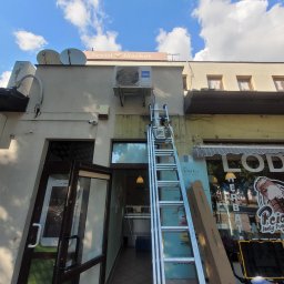 DMM Klimat Dawid Matejek - Montaż klimatyzacji Haier na fasadzie budynku Textil Market, widoczna drabina z narzędziami, błękitne niebo z chmurami w tle.