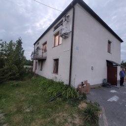 DMM Klimat Dawid Matejek - Dwupiętrowy dom z zamontowaną jednostką klimatyzacji na elewacji, widoczny balkon z czarną balustradą, kartonowe pudełka przy ścianie, mężczyzna przy wejściu do budynku.