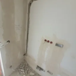 Przygotowania do instalacji w Łukowie: ściana z wyżłobieniami na kable i otwory pod gniazdka, gruz na podłodze, widoczne przewody elektryczne.