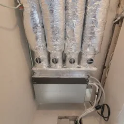 Cztery srebrne, elastyczne przewody wentylacyjne podłączone do skrzynki rozdzielczej w jasnym pomieszczeniu, z widocznymi przewodami i otworami w ścianie.