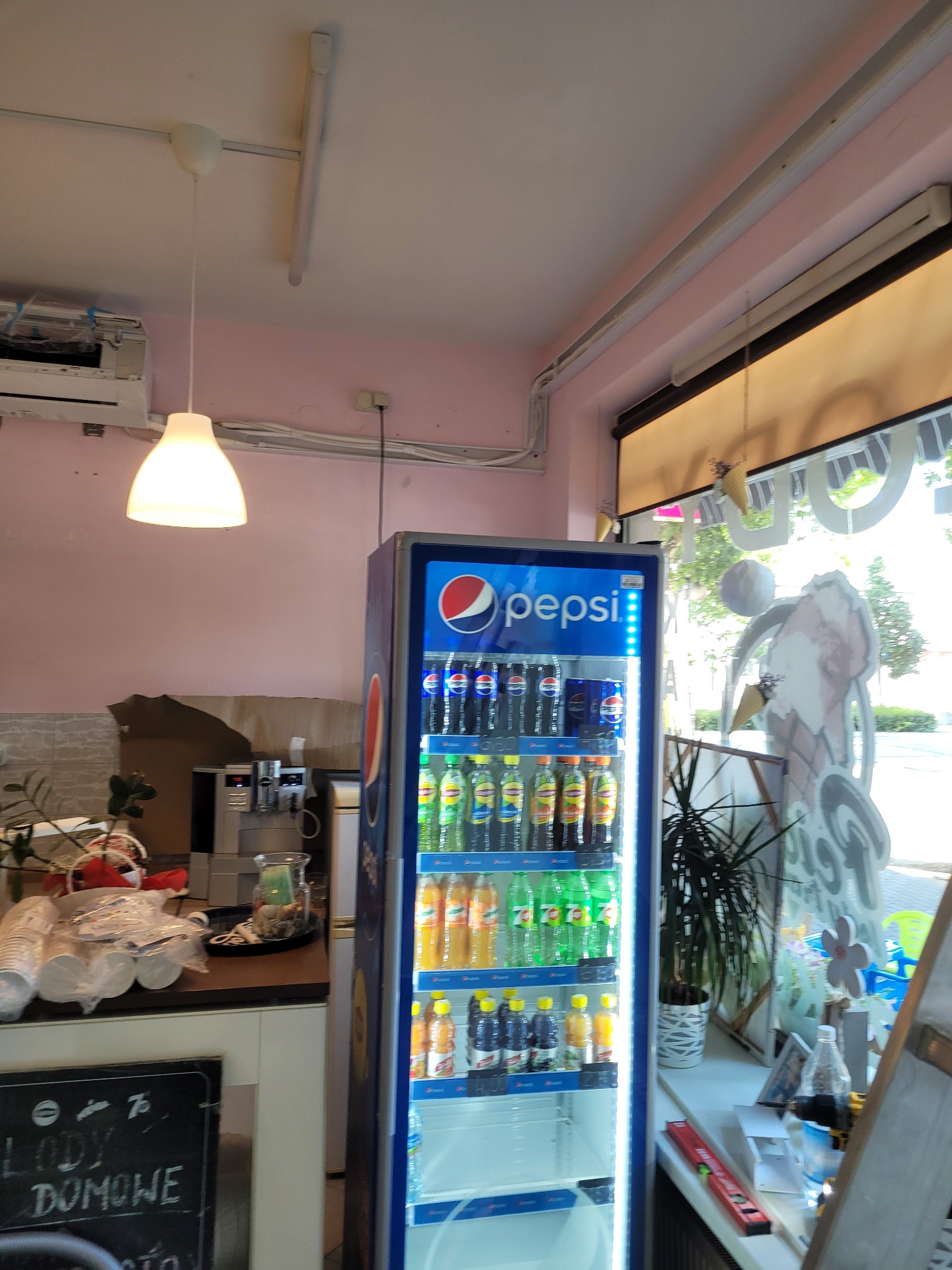 Wnętrze lokalu gastronomicznego z lodówką chłodniczą Pepsi, ekspres do kawy i tablica z napisem 'Lody Domowe'.