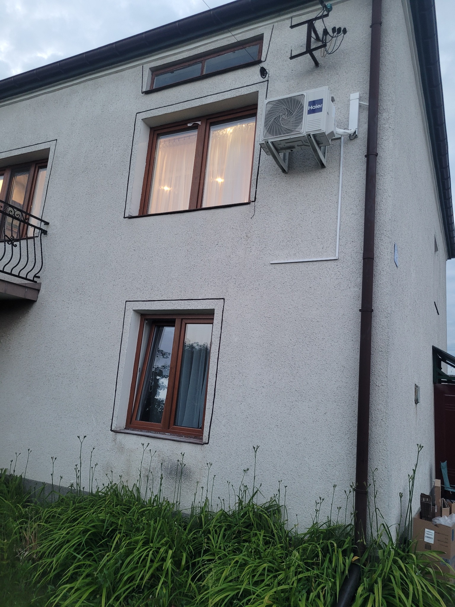 Biała jednostka zewnętrzna klimatyzacji Haier zamontowana na ścianie dwupiętrowego domu z teksturą baranka, widok z dołu na tle pochmurnego nieba.