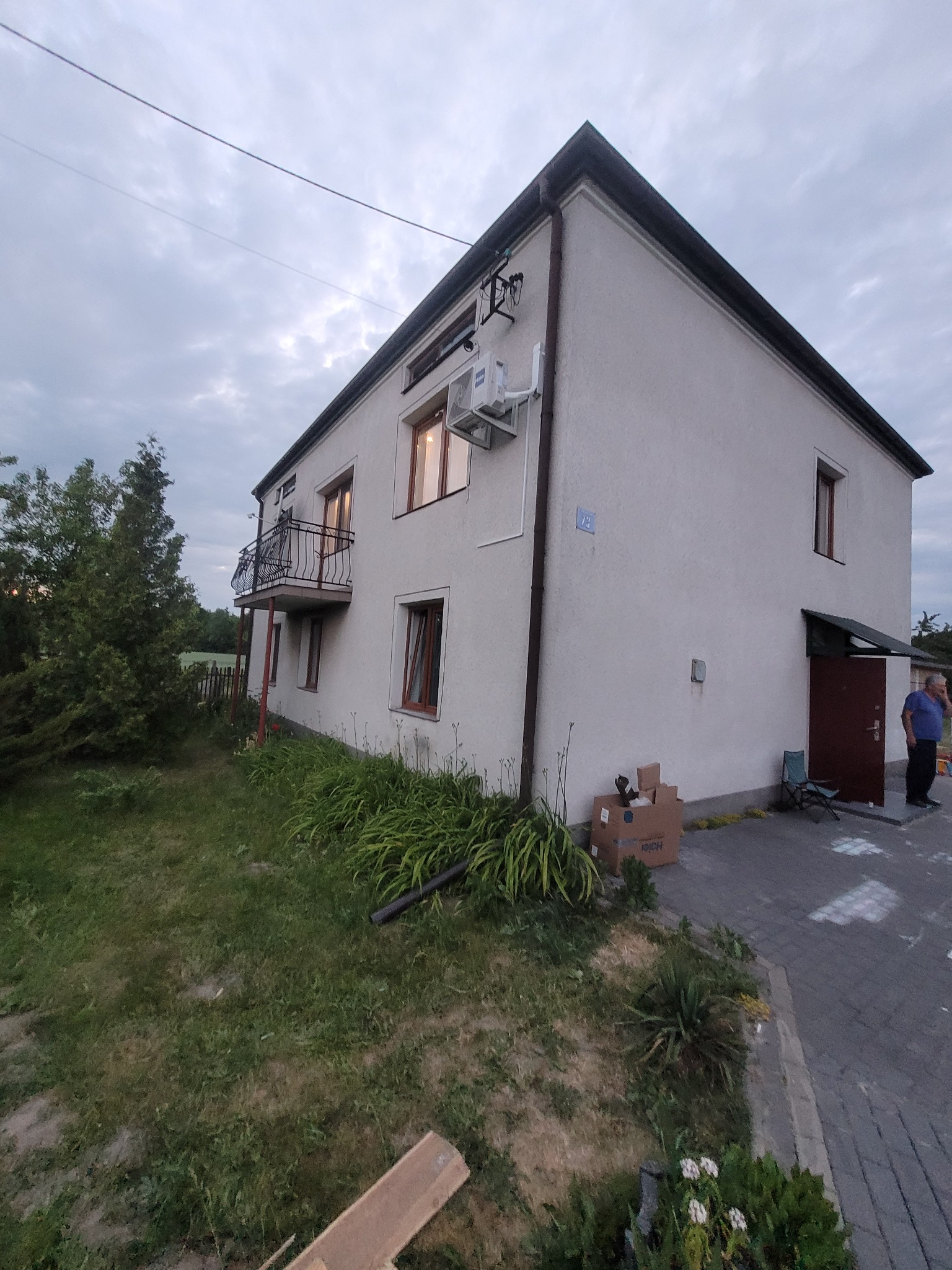 Dwupiętrowy dom z zamontowaną jednostką klimatyzacji na elewacji, widoczny balkon z czarną balustradą, kartonowe pudełka przy ścianie, mężczyzna przy wejściu do budynku.