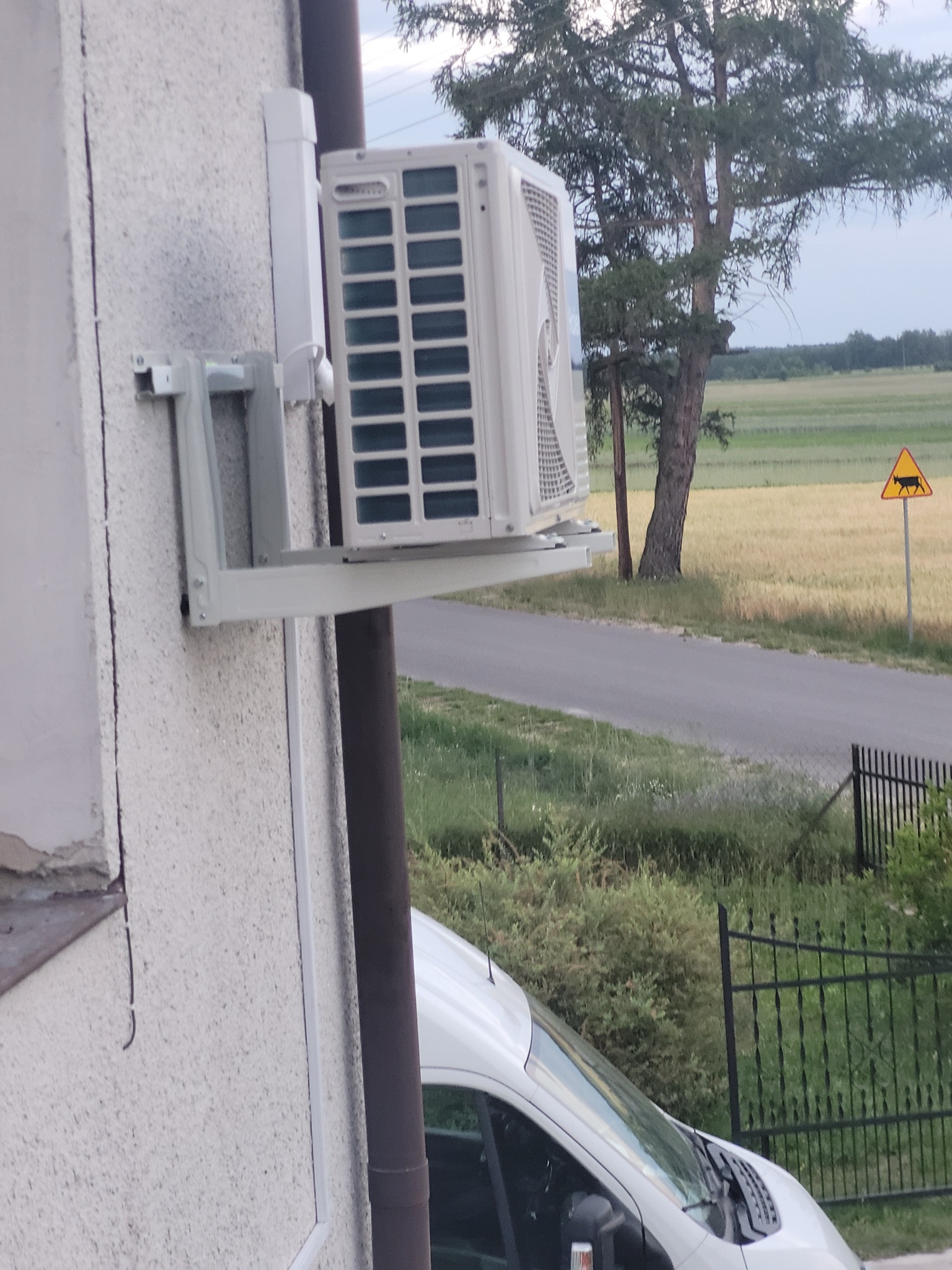 Zewnętrzna jednostka klimatyzacji zamontowana na ścianie budynku, widoczny fragment białego samochodu i znak ostrzegawczy o zwierzętach na drodze w tle.