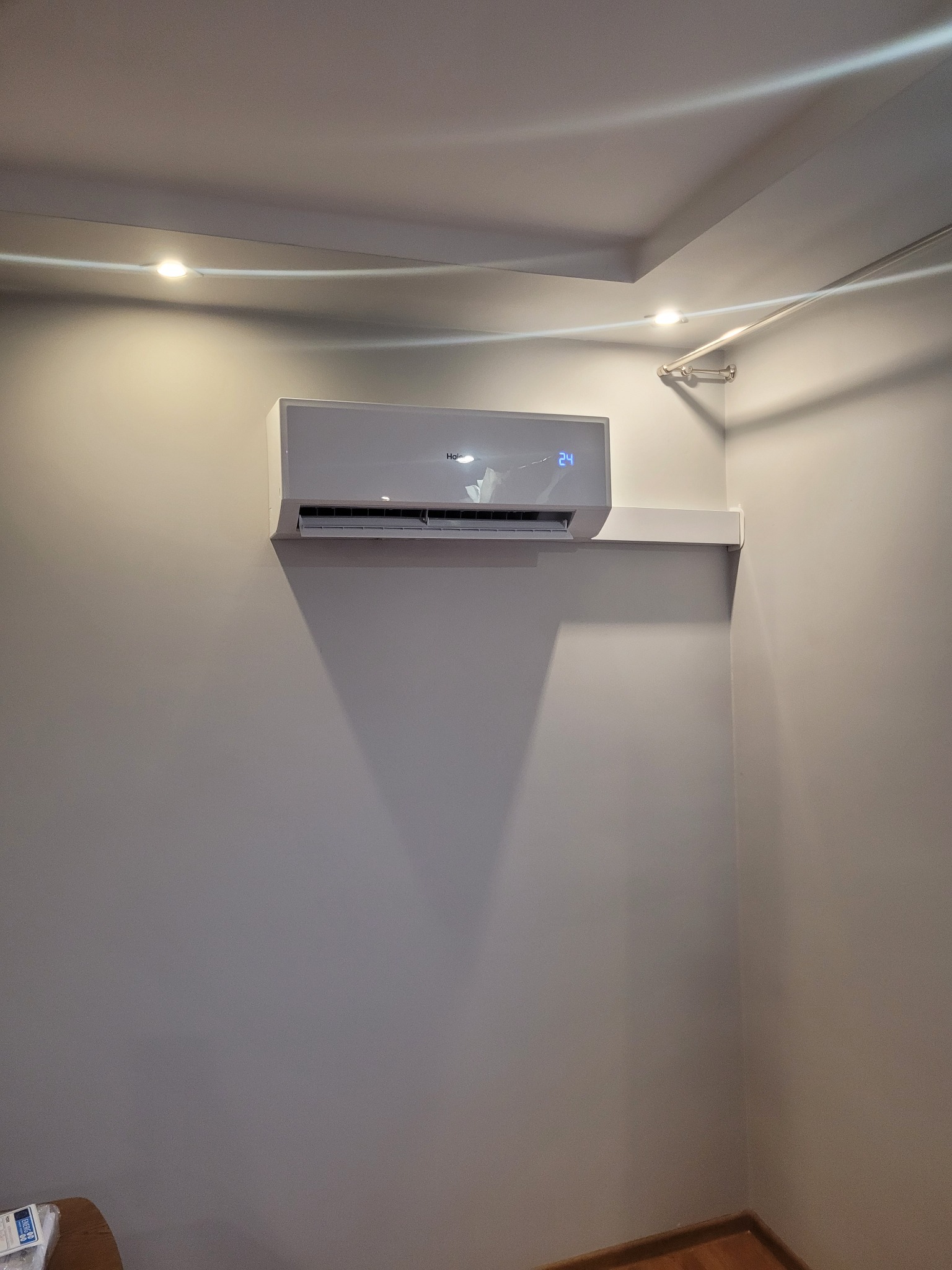 Biała klimatyzacja Haier zamontowana na ścianie w rogu pokoju, wyświetlająca temperaturę 24 stopnie, z widocznymi punktowymi lampami sufitowymi i drewnianą podłogą.