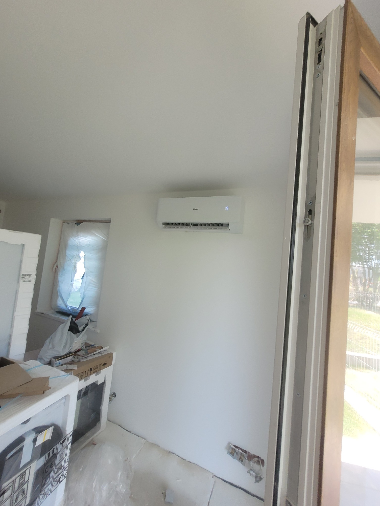 Biały klimatyzator Haier zamontowany na ścianie w surowym wnętrzu, widoczne okno zabezpieczone folią malarską i resztki po remoncie.