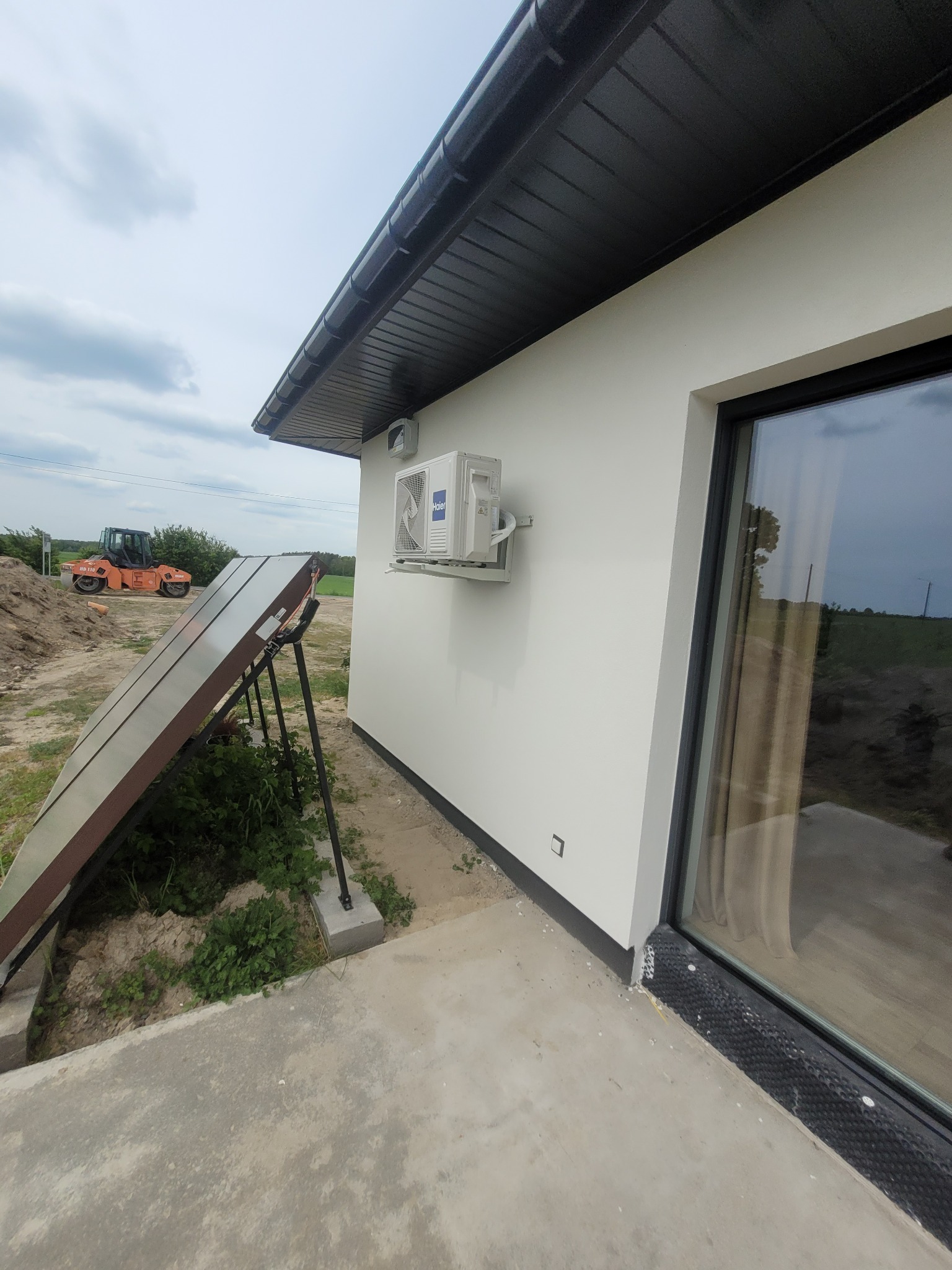 Zewnętrzna jednostka klimatyzacji na elewacji nowoczesnego domu z widocznymi panelami słonecznymi i walcem drogowym w tle.