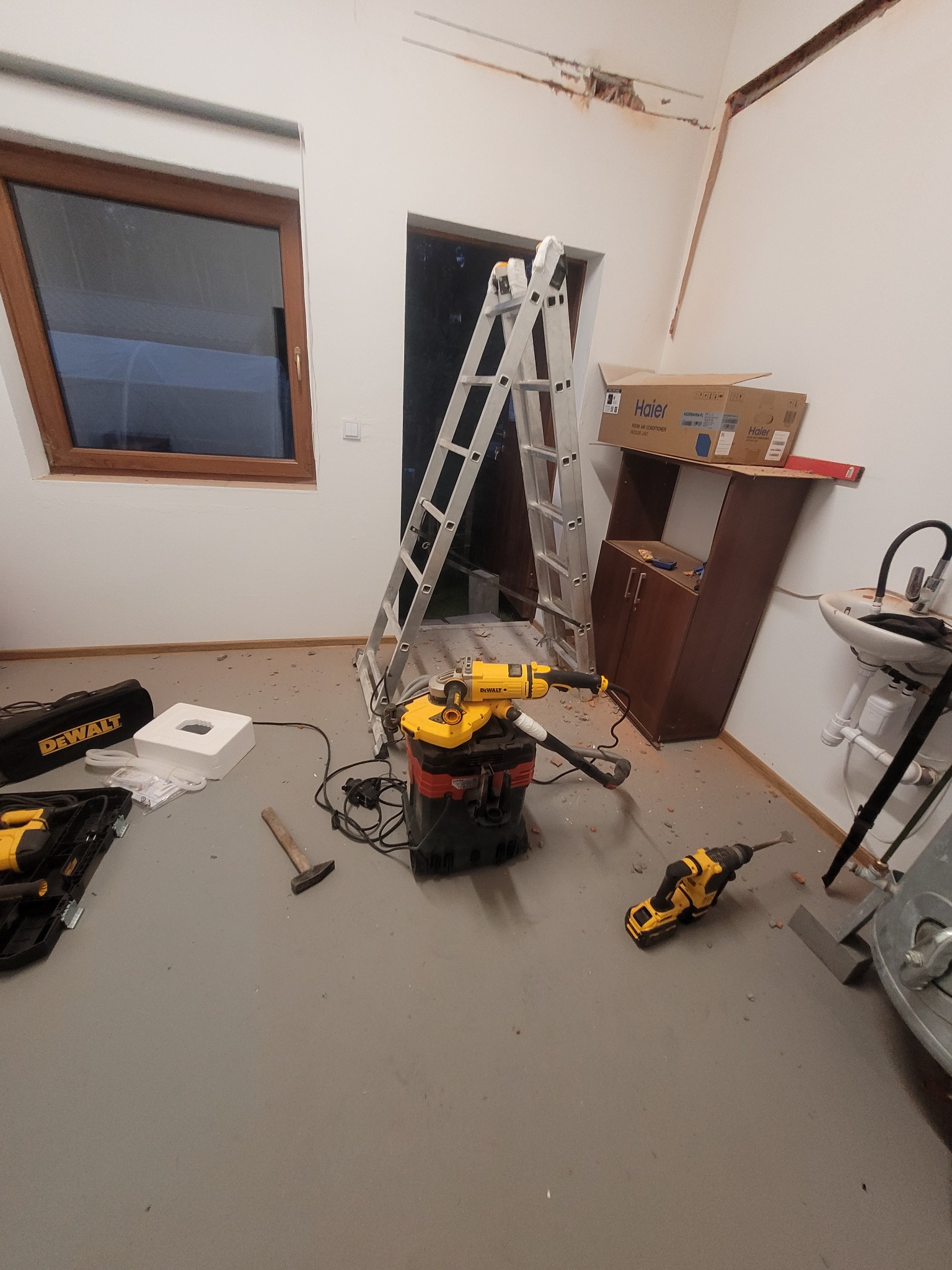 Wnętrze w trakcie remontu: drabina oparta o ścianę, narzędzia DeWalt rozłożone na podłodze, widoczne ślady prac na suficie, karton po klimatyzatorze Haier na szafce.