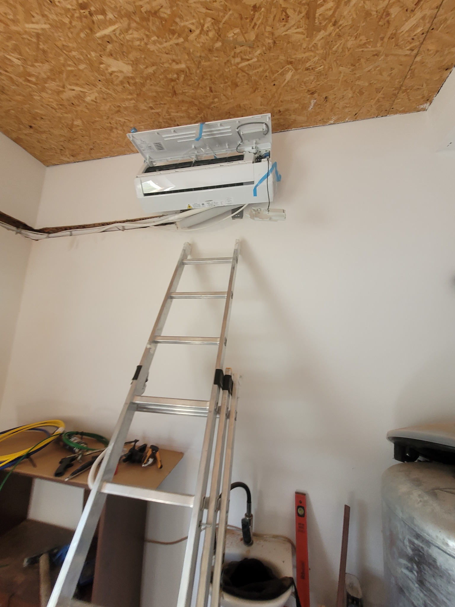 Montaż klimatyzatora split na białej ścianie w pomieszczeniu z surowym drewnianym sufitem, widoczna rozłożona drabina aluminiowa i narzędzia na półce, elementy instalacji w trakcie podłączania.