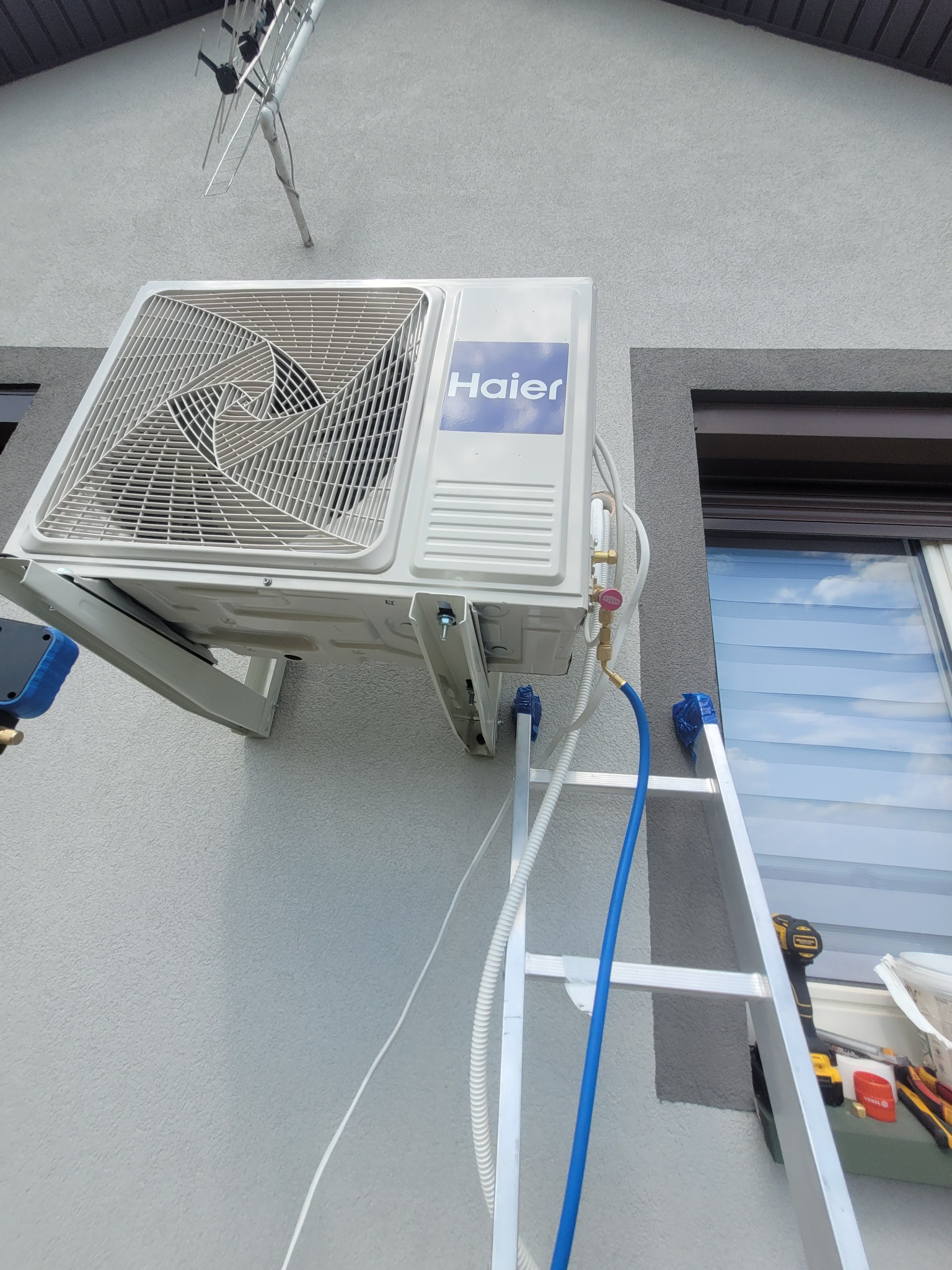 Zewnętrzna jednostka klimatyzacji Haier zamontowana na elewacji domu, widoczna drabina z narzędziami, niebieski przewód podłączony do zaworu, antena na dachu.