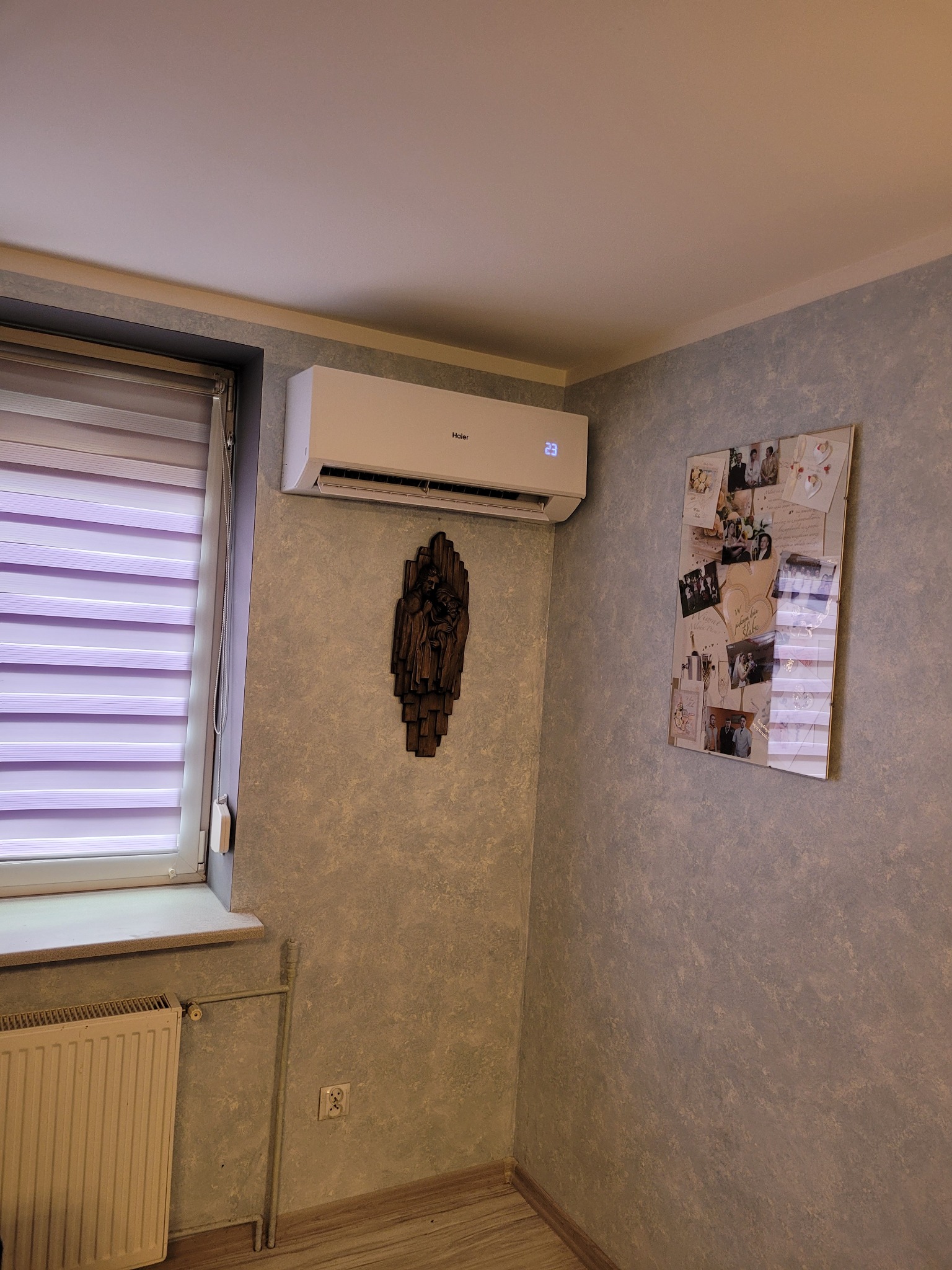 Biały klimatyzator Haier zamontowany na ścianie w pokoju z szarymi ścianami i dekoracjami, w tle okno z żaluzjami i grzejnik.