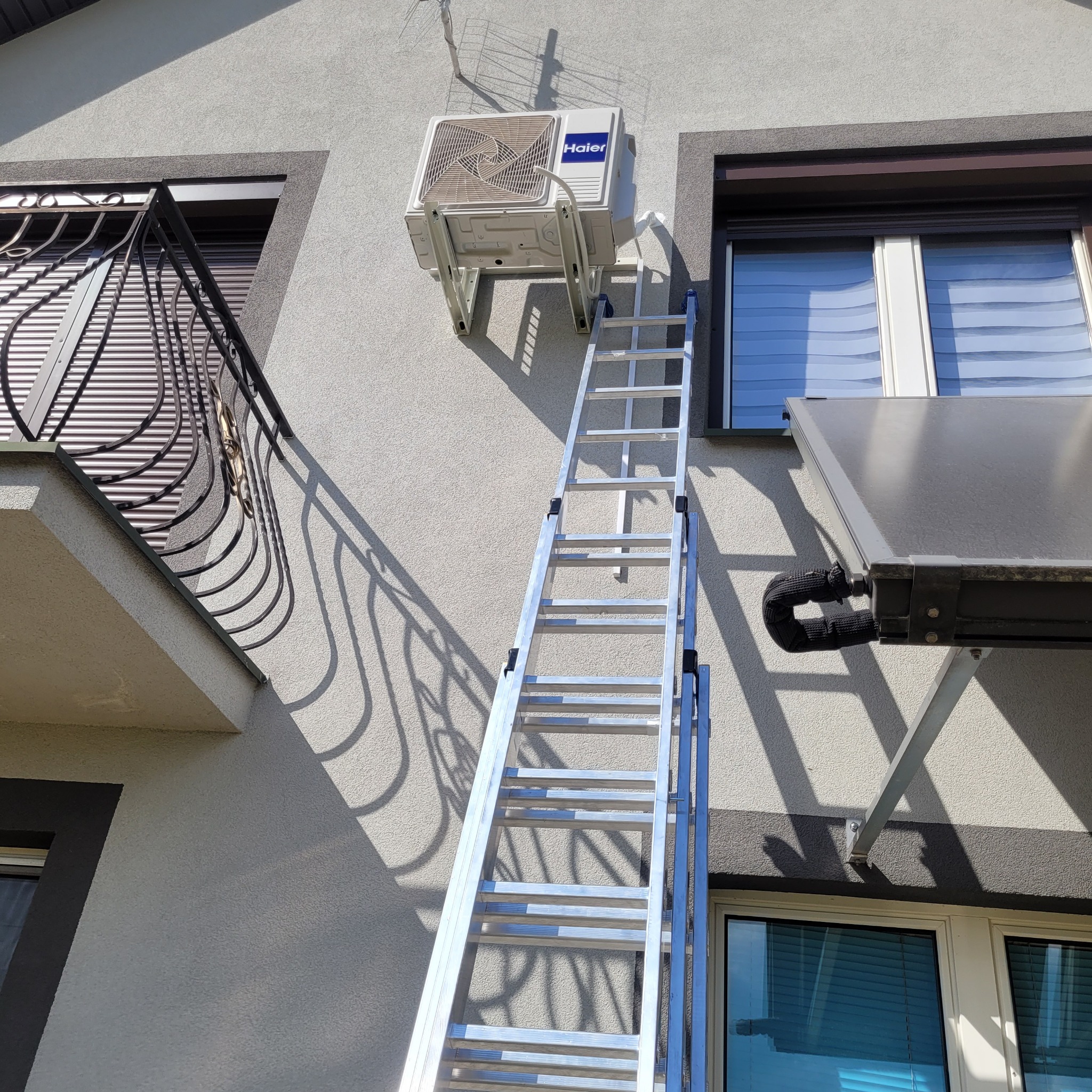 Zewnętrzna jednostka klimatyzacji Haier zamontowana na elewacji domu, dostępna za pomocą rozstawionej aluminiowej drabiny, obok okna z roletą zewnętrzną i kolektora słonecznego.