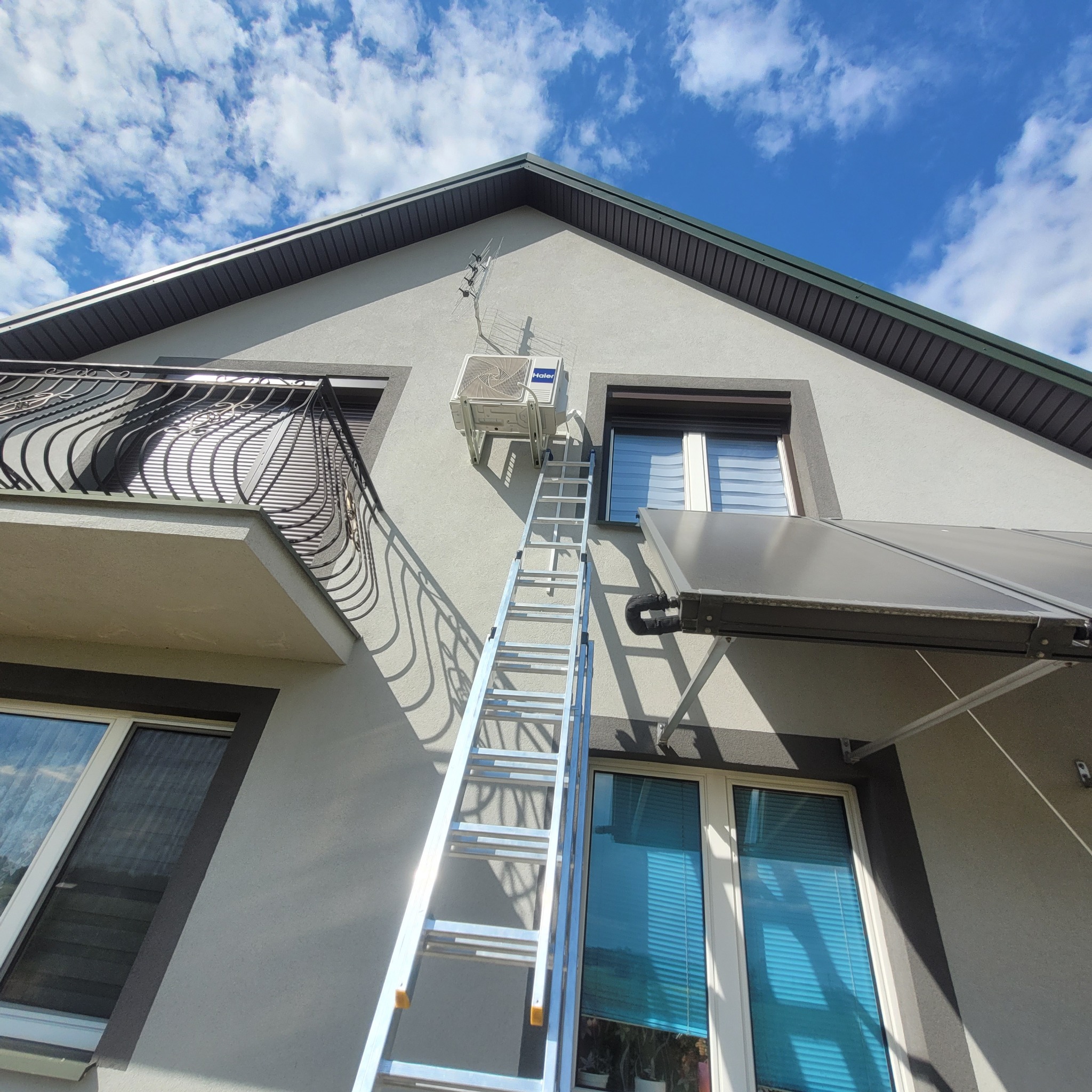 Zewnętrzny klimatyzator marki Haier zamontowany na elewacji domu, dostępny za pomocą aluminiowej drabiny rozstawnej, na tle błękitnego nieba z chmurami.