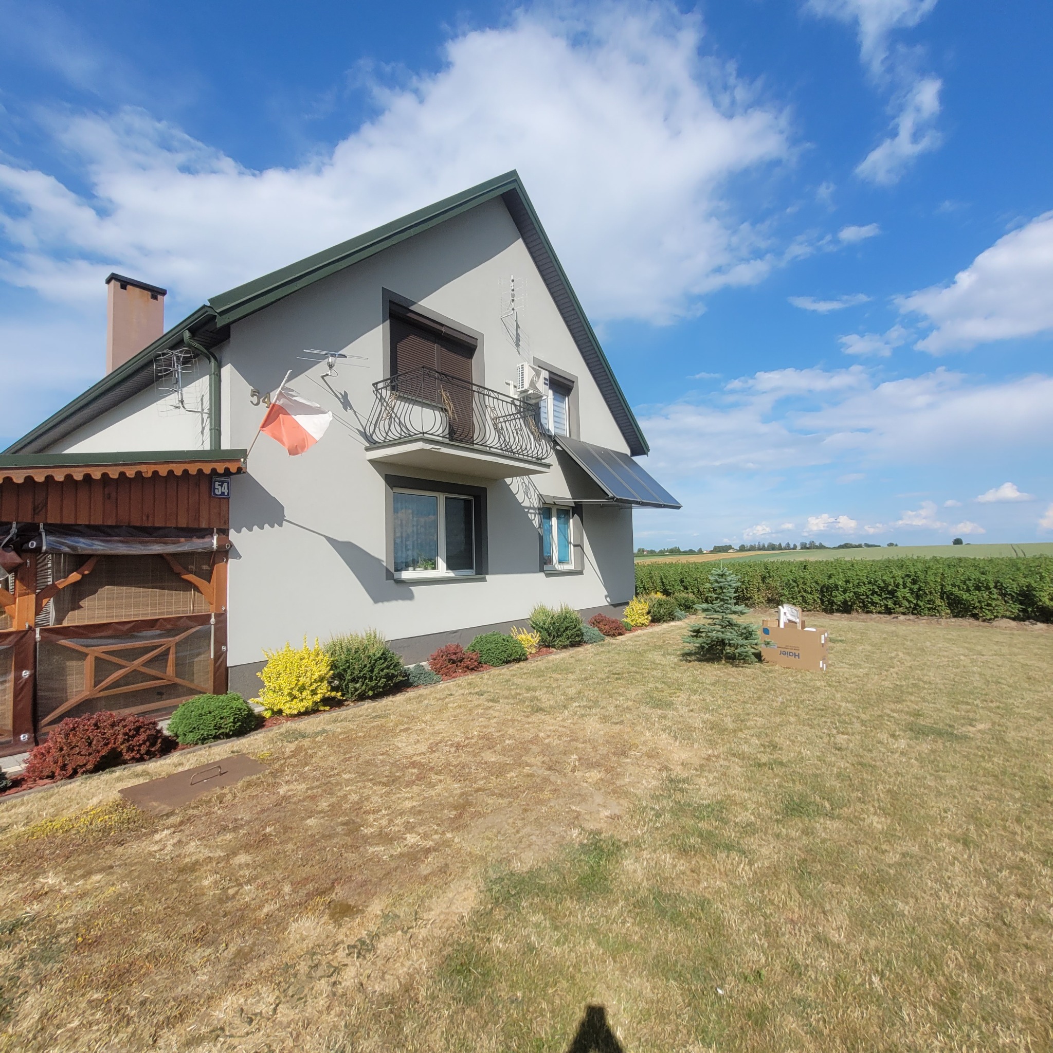 Dom jednorodzinny z klimatyzatorem na balkonie i panelem fotowoltaicznym nad oknem, na trawniku kartony z napisem Haier, w tle pole uprawne.