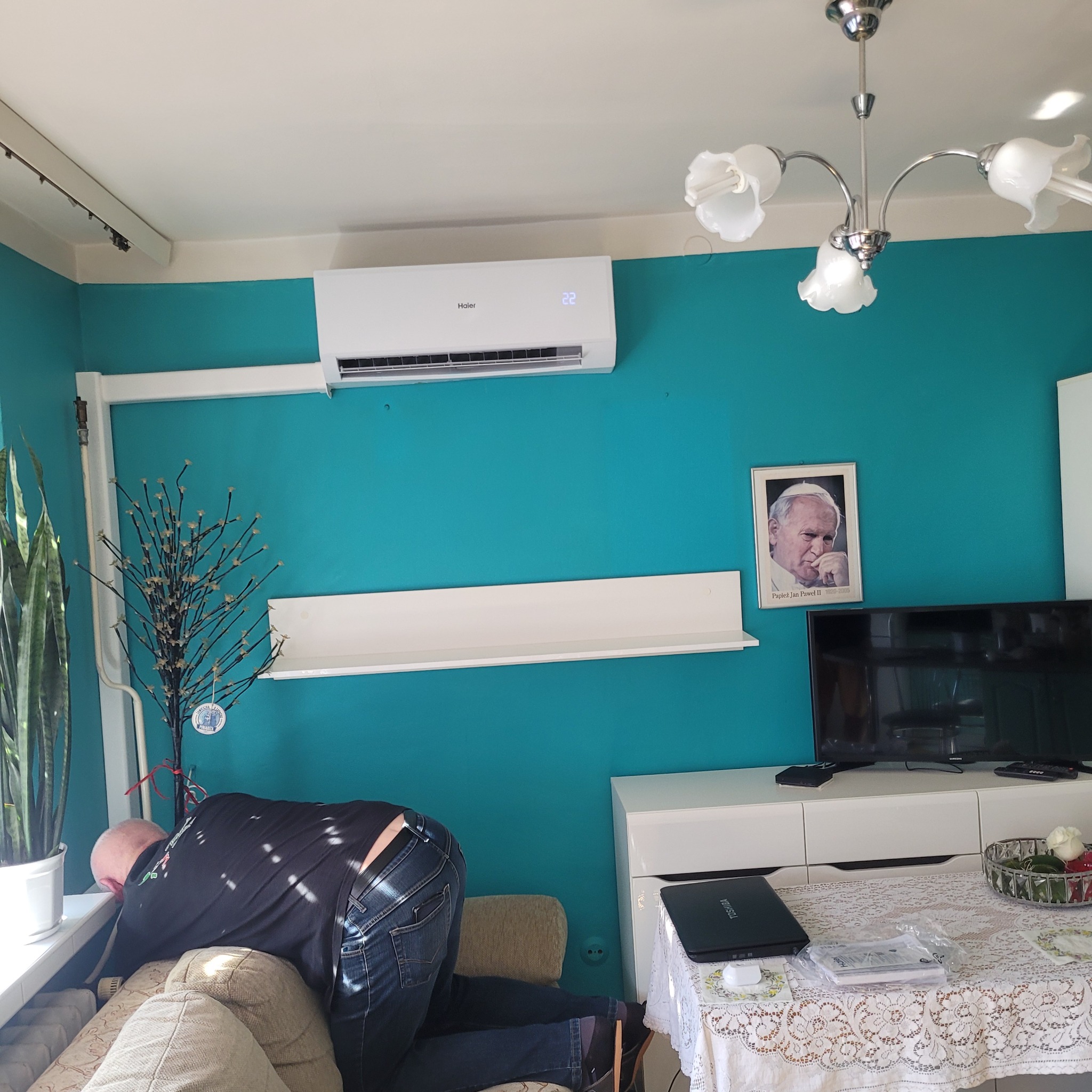Mężczyzna w trakcie montażu klimatyzatora Haier w pomieszczeniu z turkusową ścianą, widoczny portret Jana Pawła II, roślina doniczkowa i dekoracyjne gałązki.