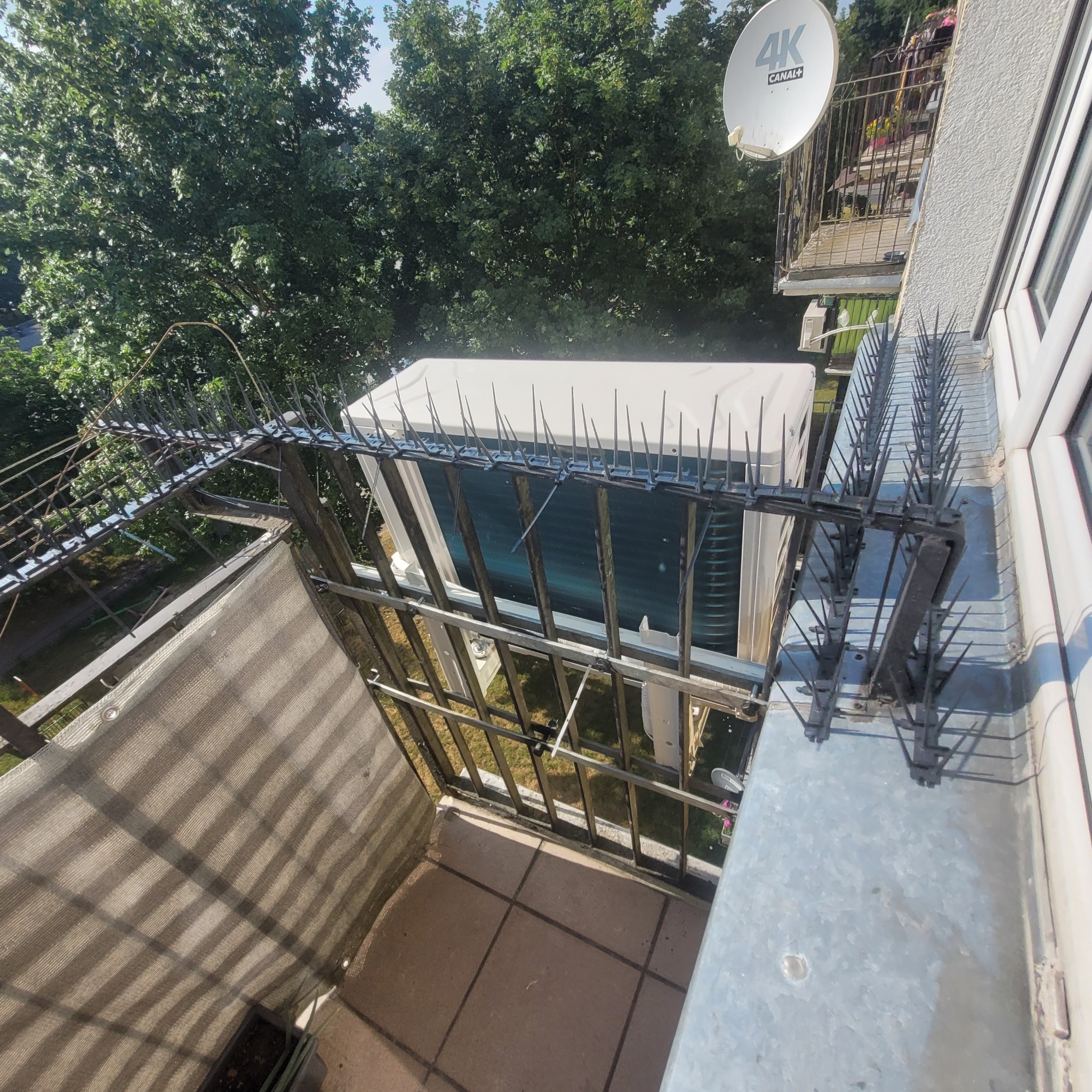 Zewnętrzna jednostka klimatyzacji na balkonie zabezpieczona kolcami przeciw ptakom, widok z góry na szary balkon z widoczną siatką cieniującą i anteną satelitarną w tle.