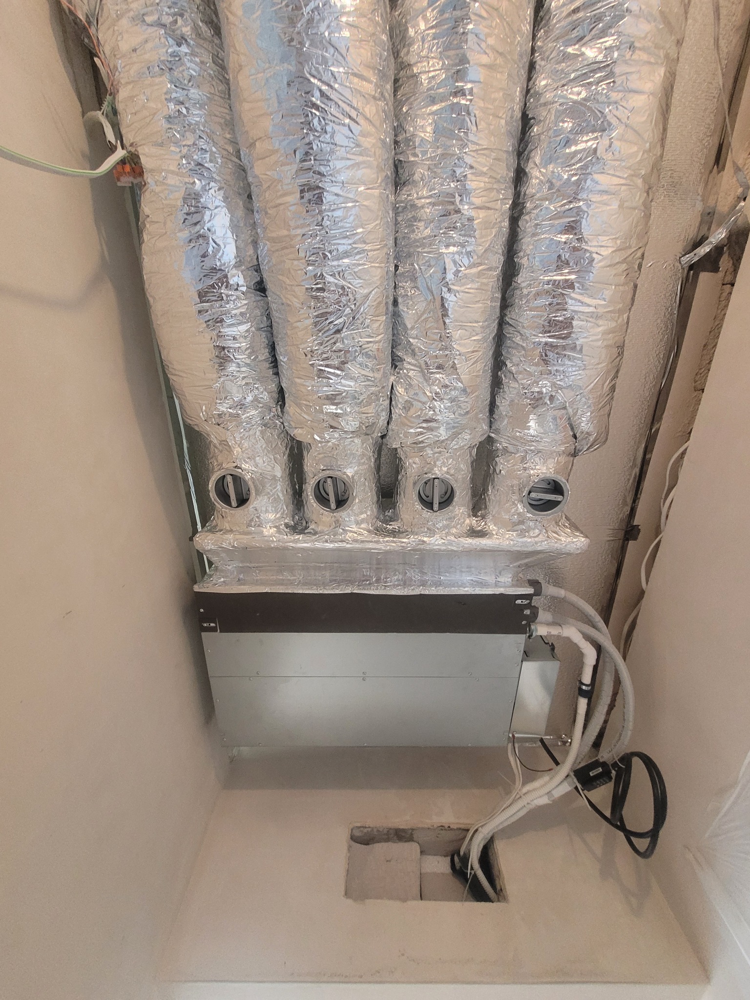 Cztery srebrne, elastyczne przewody wentylacyjne podłączone do skrzynki rozdzielczej w jasnym pomieszczeniu, z widocznymi przewodami i otworami w ścianie.