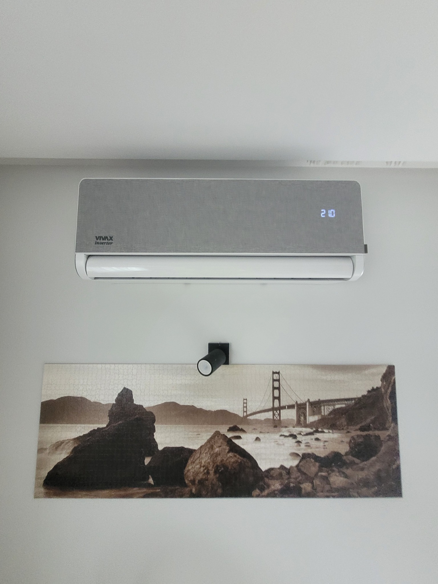 Szary klimatyzator VIVAX Inverter na białej ścianie nad obrazem z motywem Golden Gate. Widoczny czarny reflektor punktowy.