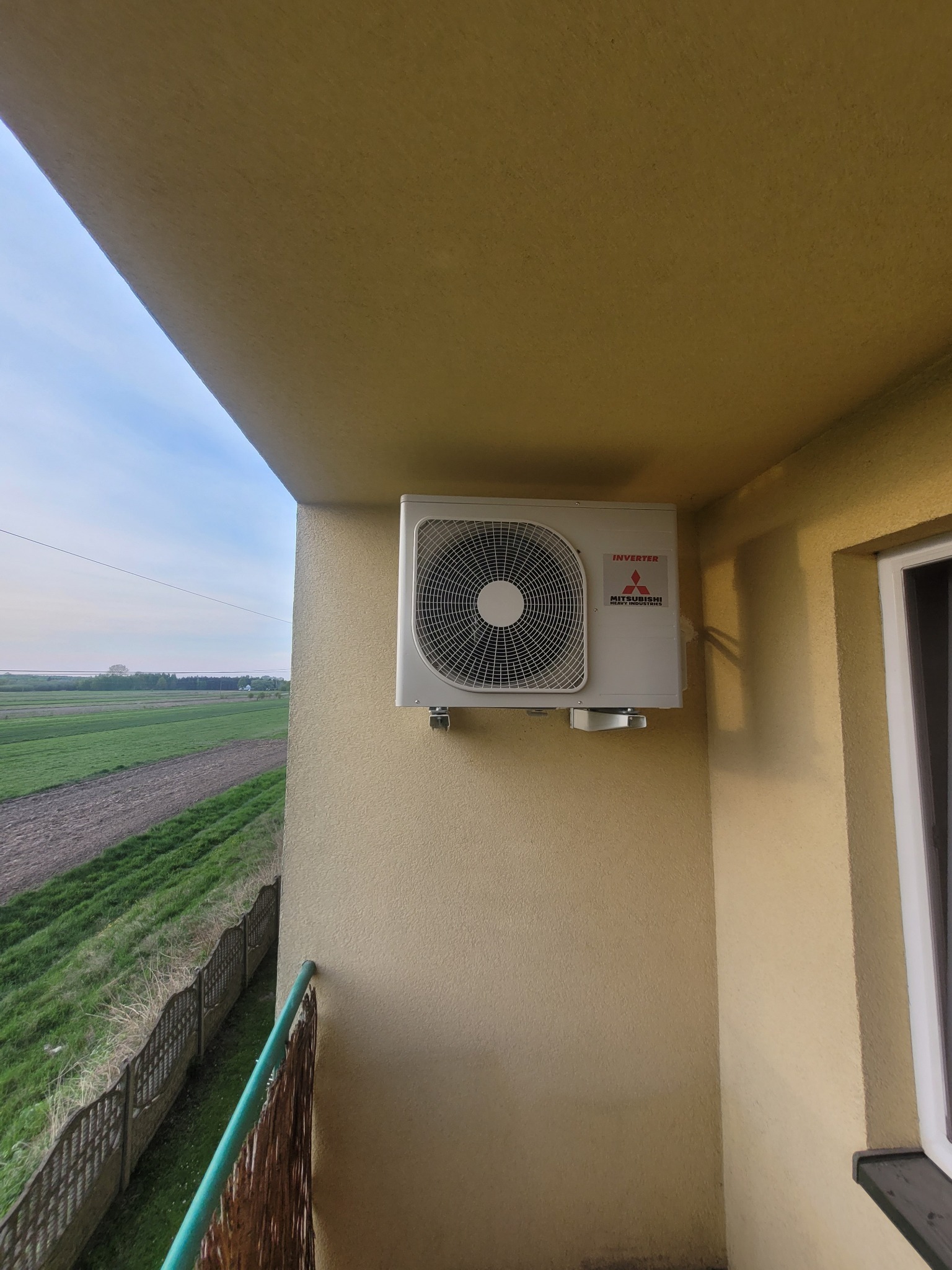 Klimatyzator Mitsubishi Inverter zamontowany na balkonie z widokiem na pola. Jasna elewacja budynku, błękitne niebo w tle, zielona balustrada.