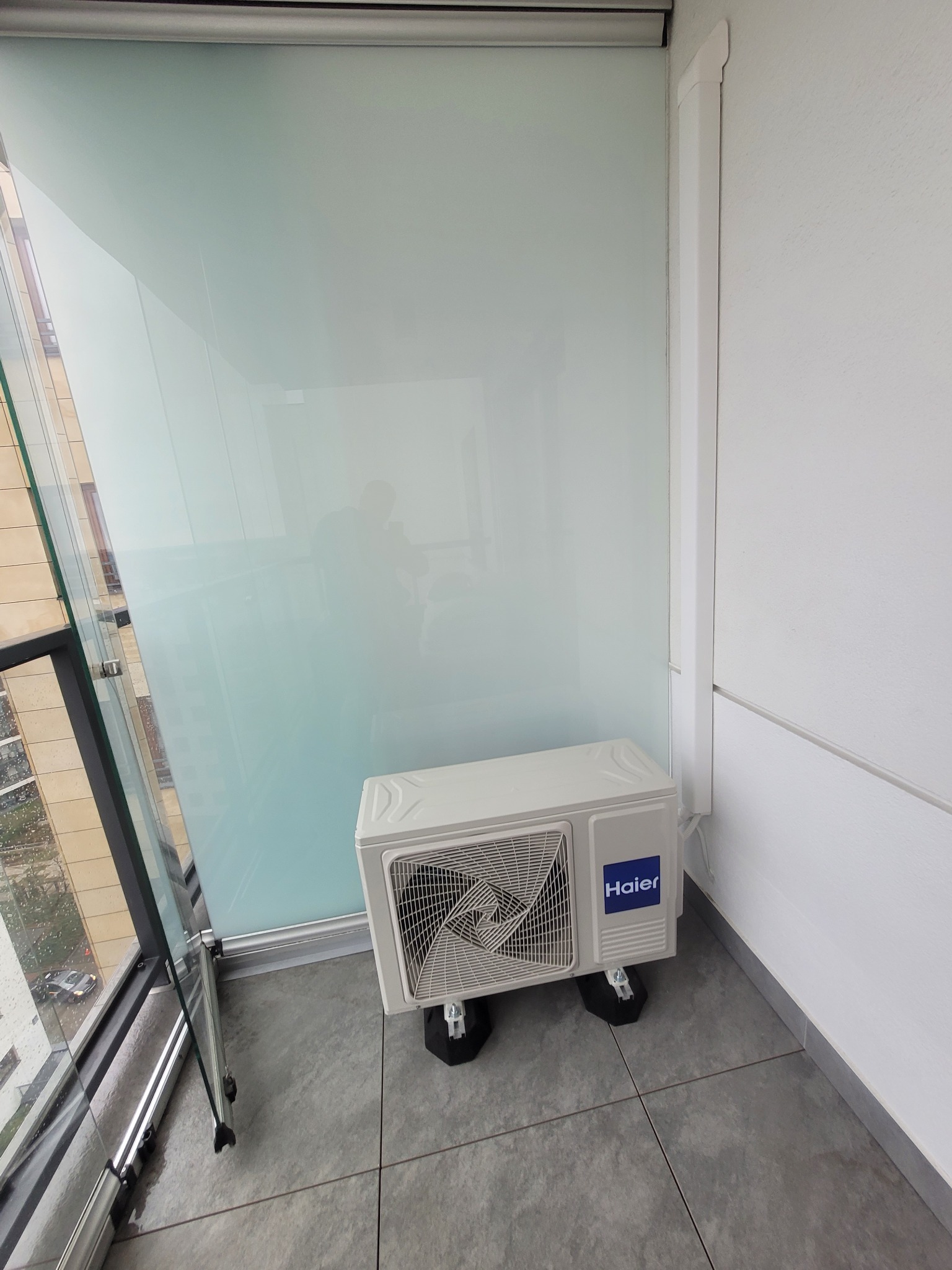Biała jednostka zewnętrzna klimatyzacji Haier na balkonie w Łukowie, zamontowana na czarnych podkładkach antywibracyjnych, widok przez przeszkloną balustradę.