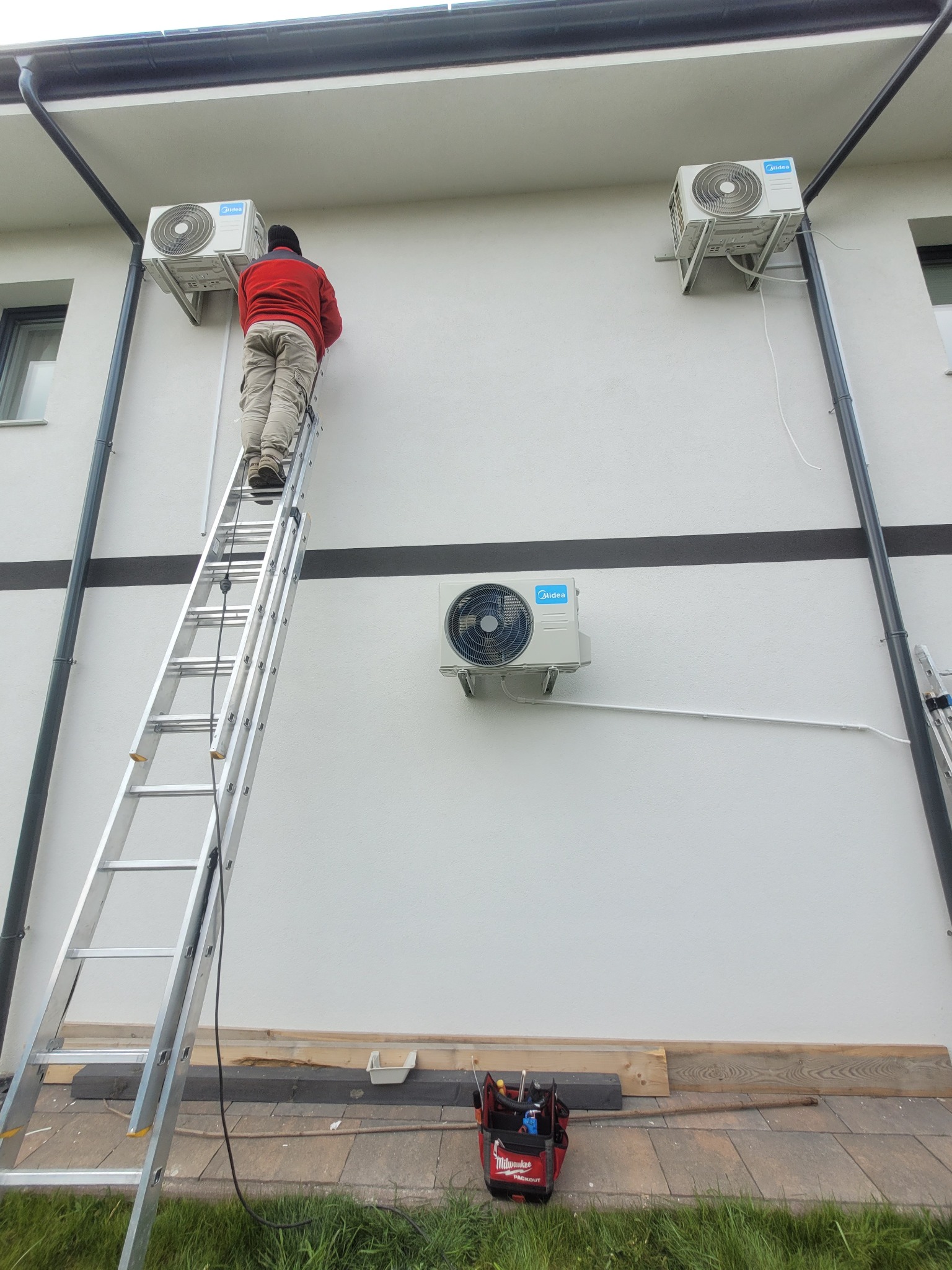 Montaż klimatyzacji Midea na elewacji domu. Instalator na drabinie przy urządzeniu, widoczna skrzynka z narzędziami. Dwa zamontowane klimatyzatory i jeden w trakcie montażu.