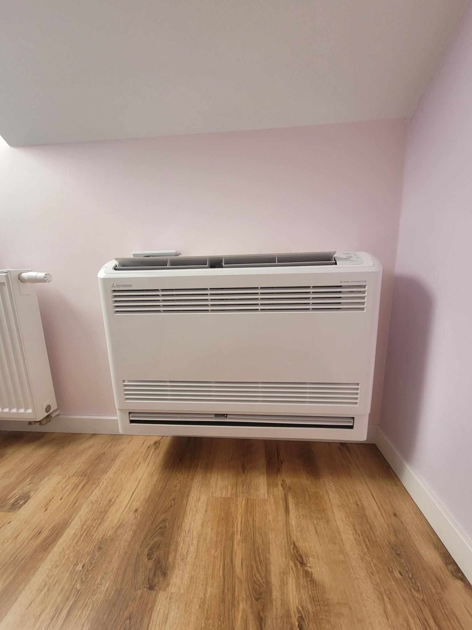 Biała, przypodłogowa klimatyzacja Mitsubishi Hyper Inverter na tle różowej ściany i drewnianej podłogi. Obok grzejnik. Widok z przodu, lekki kąt.