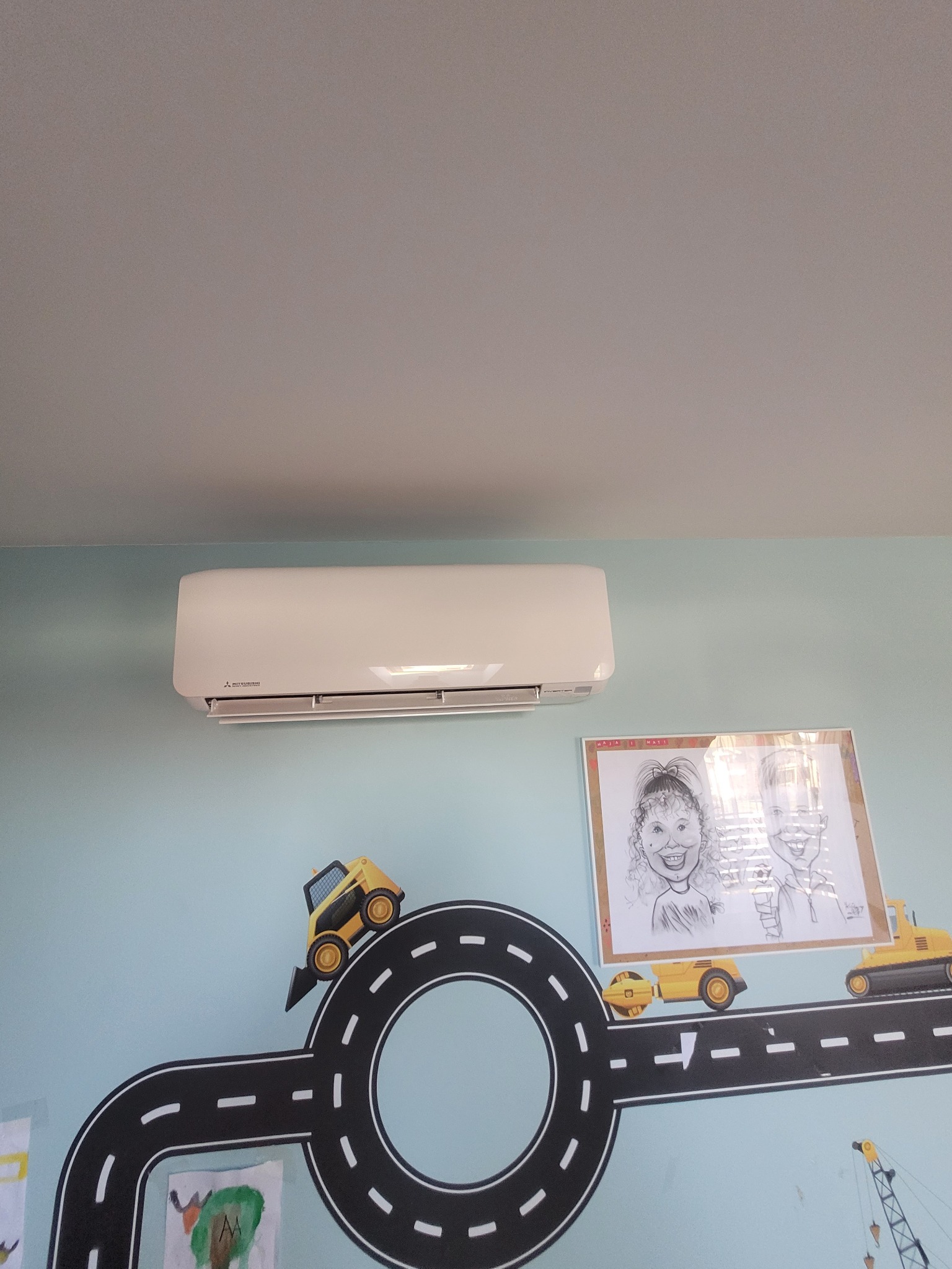 Biały klimatyzator Mitsubishi na błękitnej ścianie pokoju dziecięcego z dekoracjami drogowymi i rysunkami. Widok z dołu.