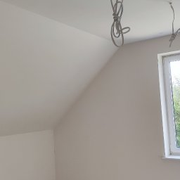 kamil osiński - Poddasze po gładzi, ściany i skos sufitu pomalowane na biało. Widoczne kable oświetleniowe i okno z widokiem na zieleń. W rogu lampa doświetlająca.